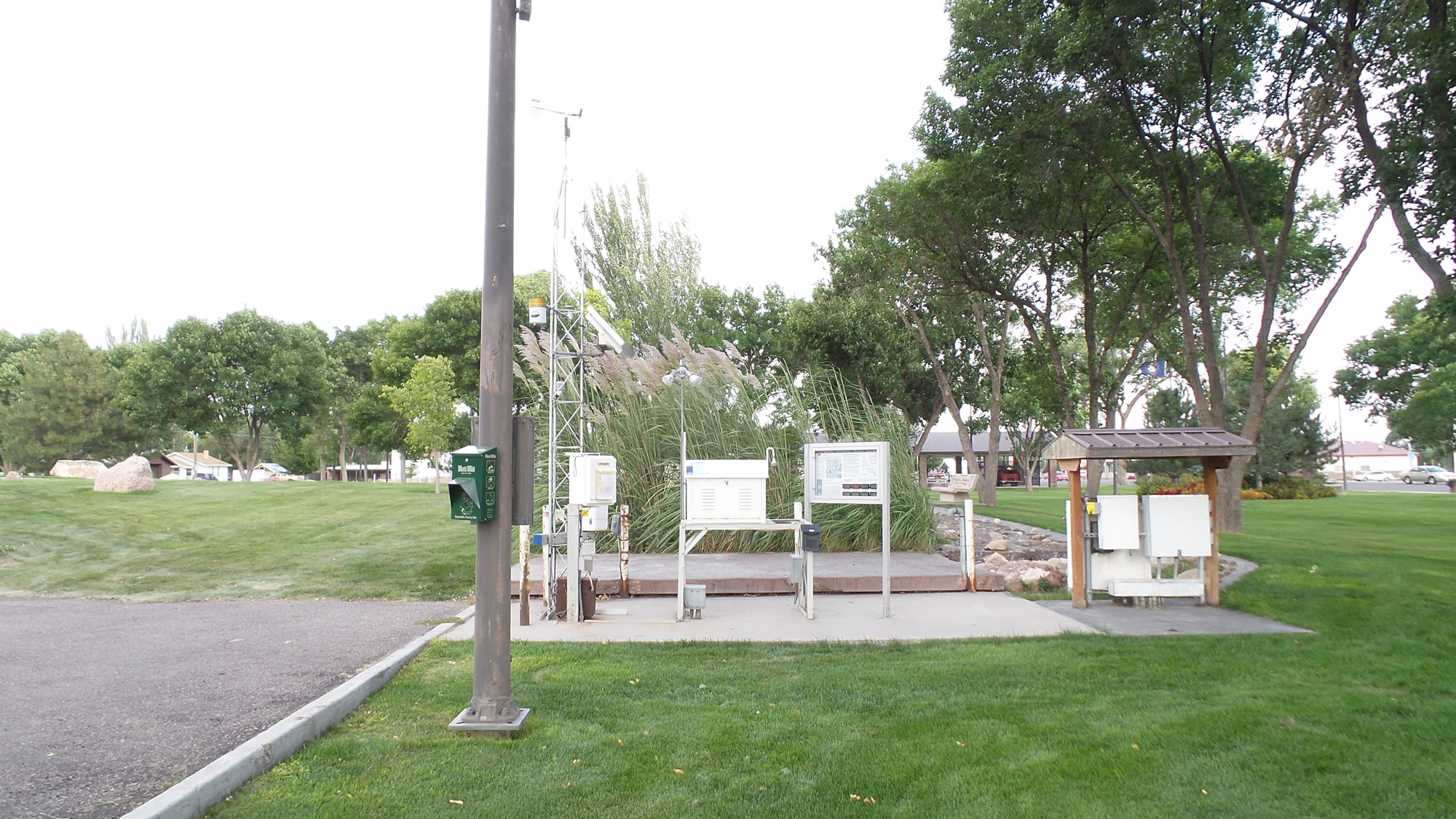 Weather monitoring station with various instruments and informational signs, surrounded by grass, trees, and a small shelter in a park setting.