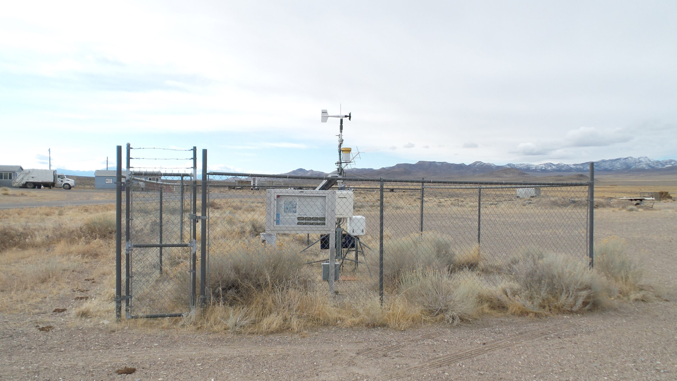 A fenced weather monitoring station stands in a dry, open landscape with mountains in the background and some buildings visible to the left.