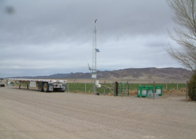 A weather monitoring station with sensors and antenna stands next to a fence and a parked flatbed trailer on a dirt road, with mountains visible in the background.