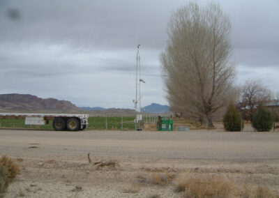 A flatbed trailer is parked near a dirt road with a tall leafless tree, a weather instrument tower, and green fields in the background under an overcast sky.