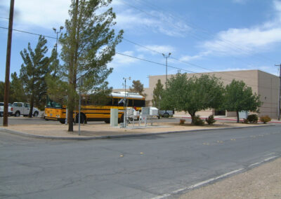 A yellow school bus is parked on a street near a beige building, with trees and utility boxes nearby under a partly cloudy sky.