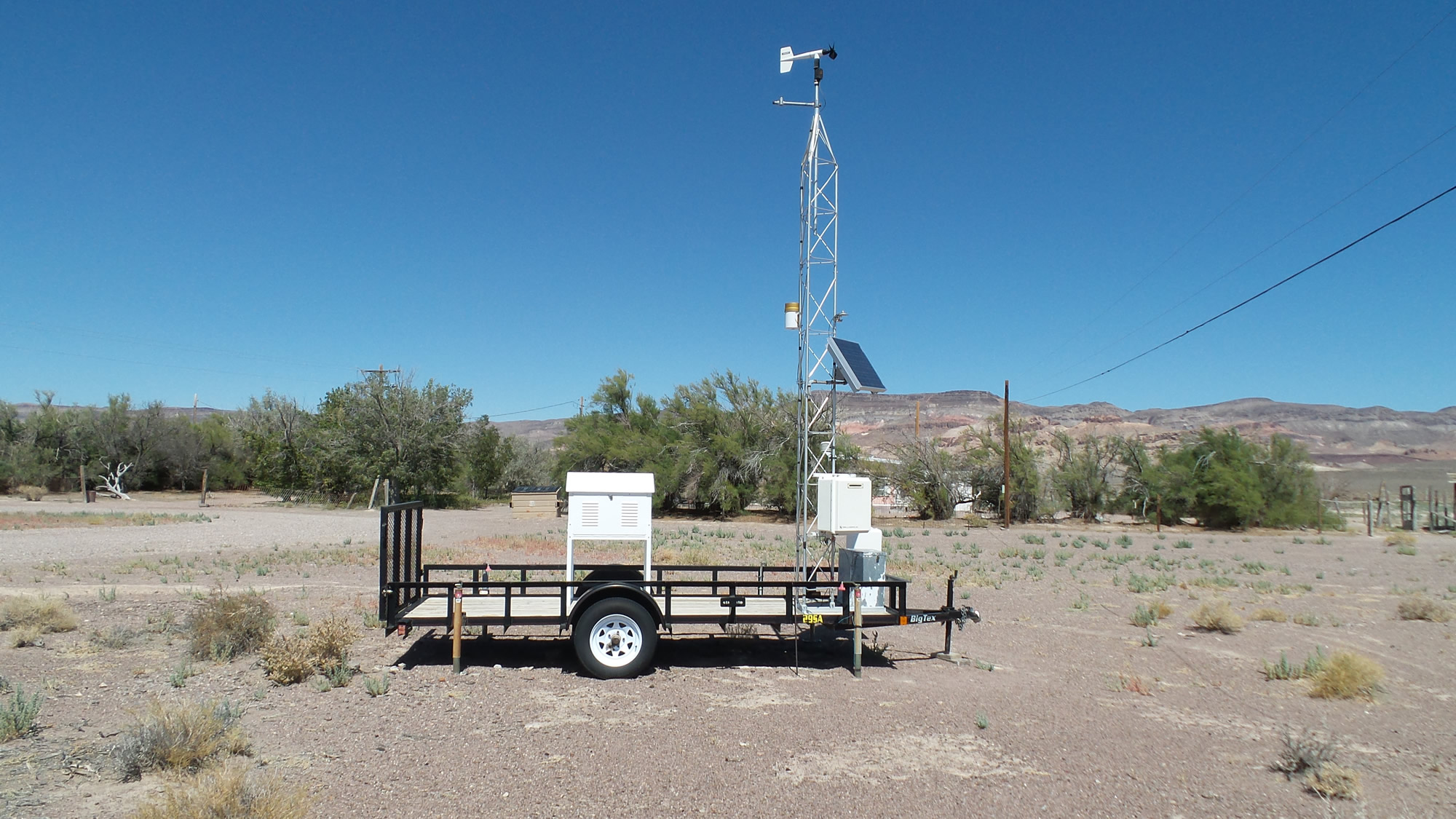 A portable weather station on a trailer features sensors, a solar panel, and a wind vane in a dry, open landscape with sparse vegetation and distant hills.