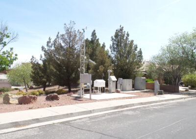 An air quality monitoring station with various instruments is set up near a suburban street, surrounded by trees and landscaping rocks.