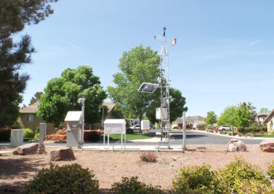 A weather station with various instruments and a solar panel is set up in a landscaped suburban area with houses and trees in the background.