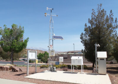 A weather station with various meteorological instruments and sensors is set up on a concrete pad, surrounded by trees and buildings under a clear sky.