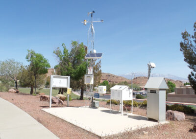 A roadside weather monitoring station with sensors, solar panel, and instrumentation, surrounded by trees and a desert landscape under a clear sky.