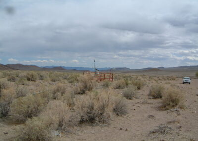 A fenced weather station stands in a dry, shrub-covered desert landscape under a cloudy sky; a white vehicle is parked at the right edge.