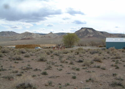 A rural landscape with dry, sparse vegetation in the foreground, a few buildings, hay bales, and a tree in the midground, and mountains in the background under a cloudy sky.