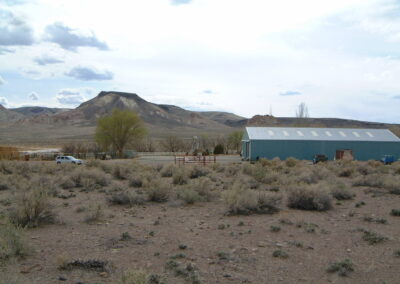 A blue metal building and a parked car stand near fenced areas in a dry, scrubby landscape with hills and sparse trees in the background.