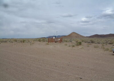 A small fenced weather station is situated in a dry, rocky desert landscape with scattered shrubs and distant hills under a cloudy sky.