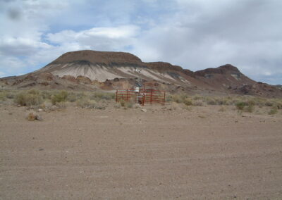 A fenced grave is situated in a dry, rocky desert landscape with sparse vegetation and hills in the background under a cloudy sky.