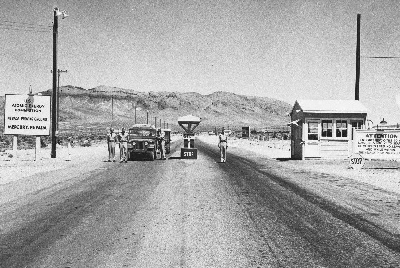 Black and white photo of a checkpoint at the Nevada Proving Ground with guards, a stop barrier, vehicles, and signs, including one for the Atomic Energy Commission.