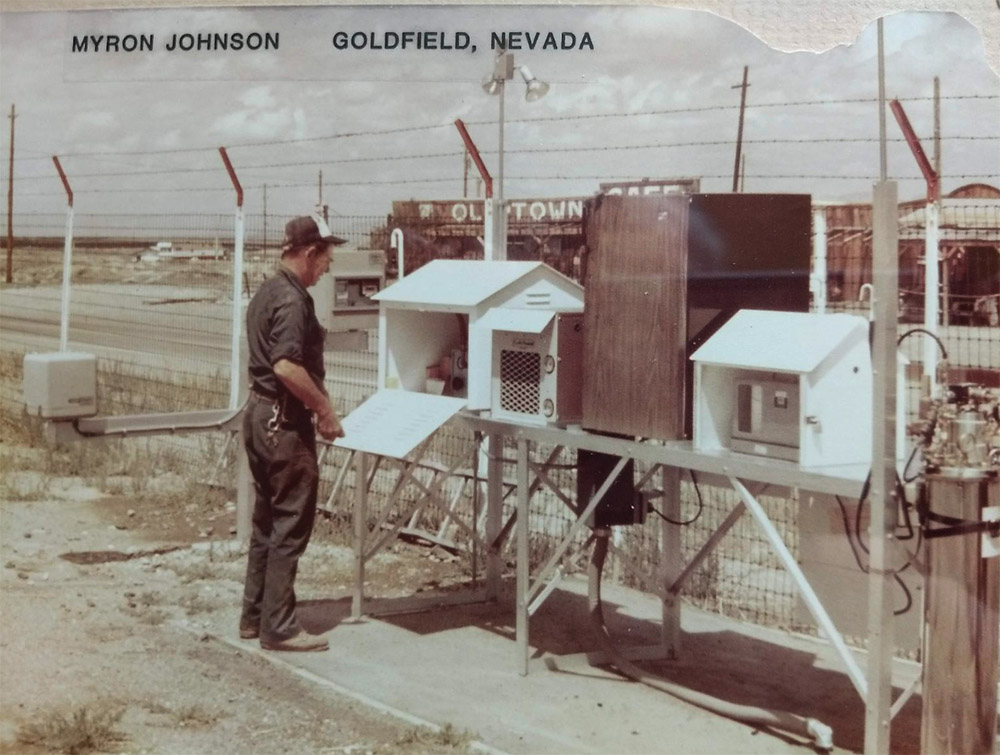 A man stands at an outdoor weather monitoring station in Goldfield, Nevada, reading equipment and data under partly cloudy skies.