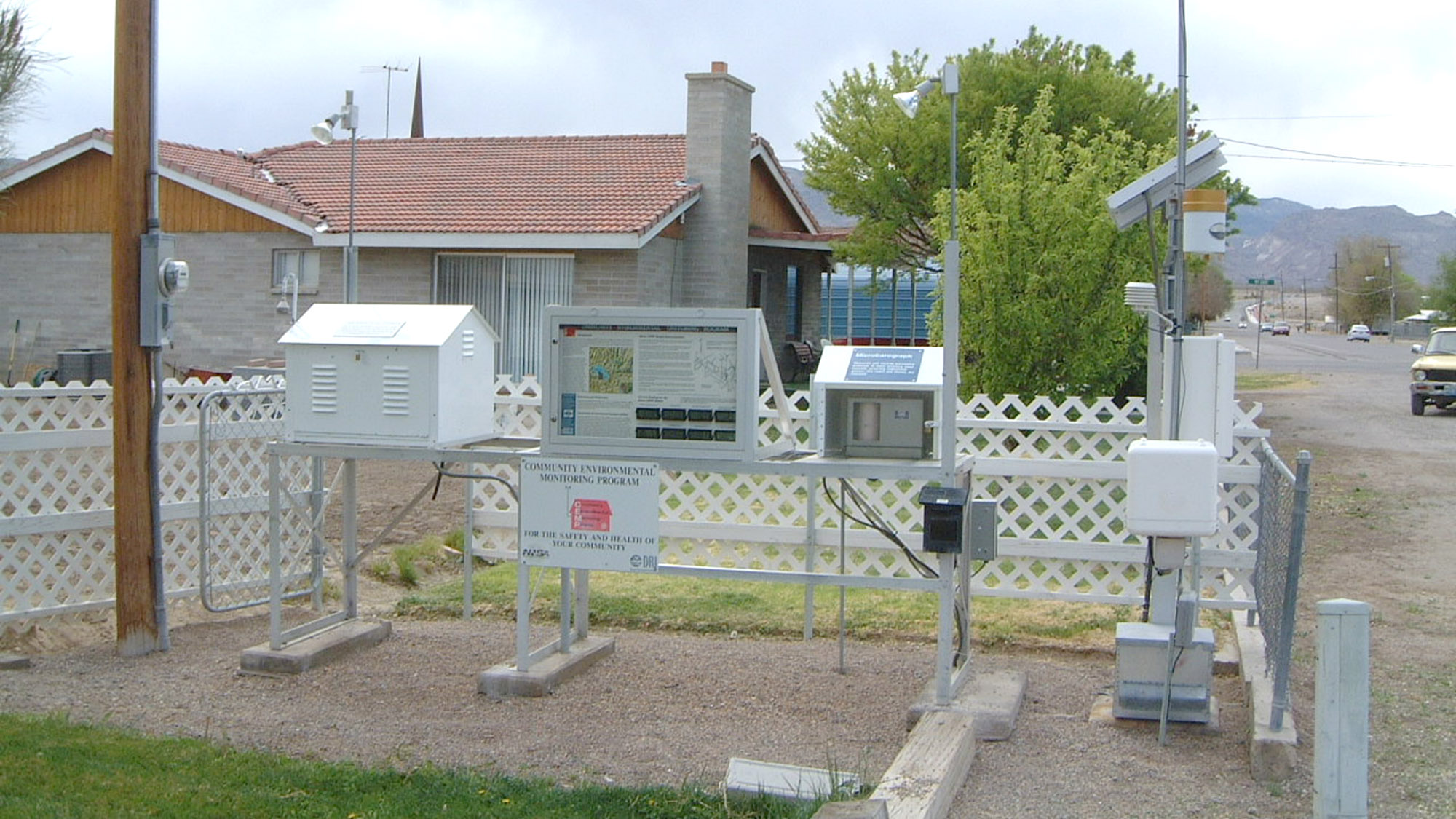 A weather monitoring station with instruments, data displays, and sensor equipment set up on a gravel area in front of a house with a white fence.