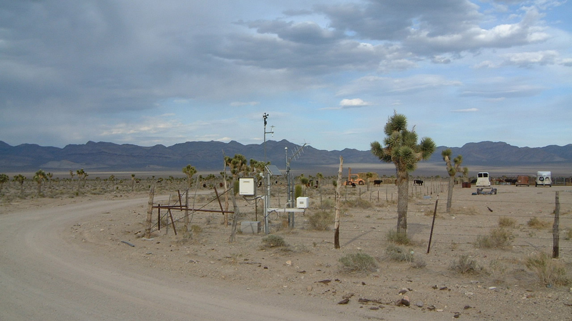 A weather monitoring station with equipment and fencing stands in a dry, desert landscape with Joshua trees and distant mountains under a cloudy sky.