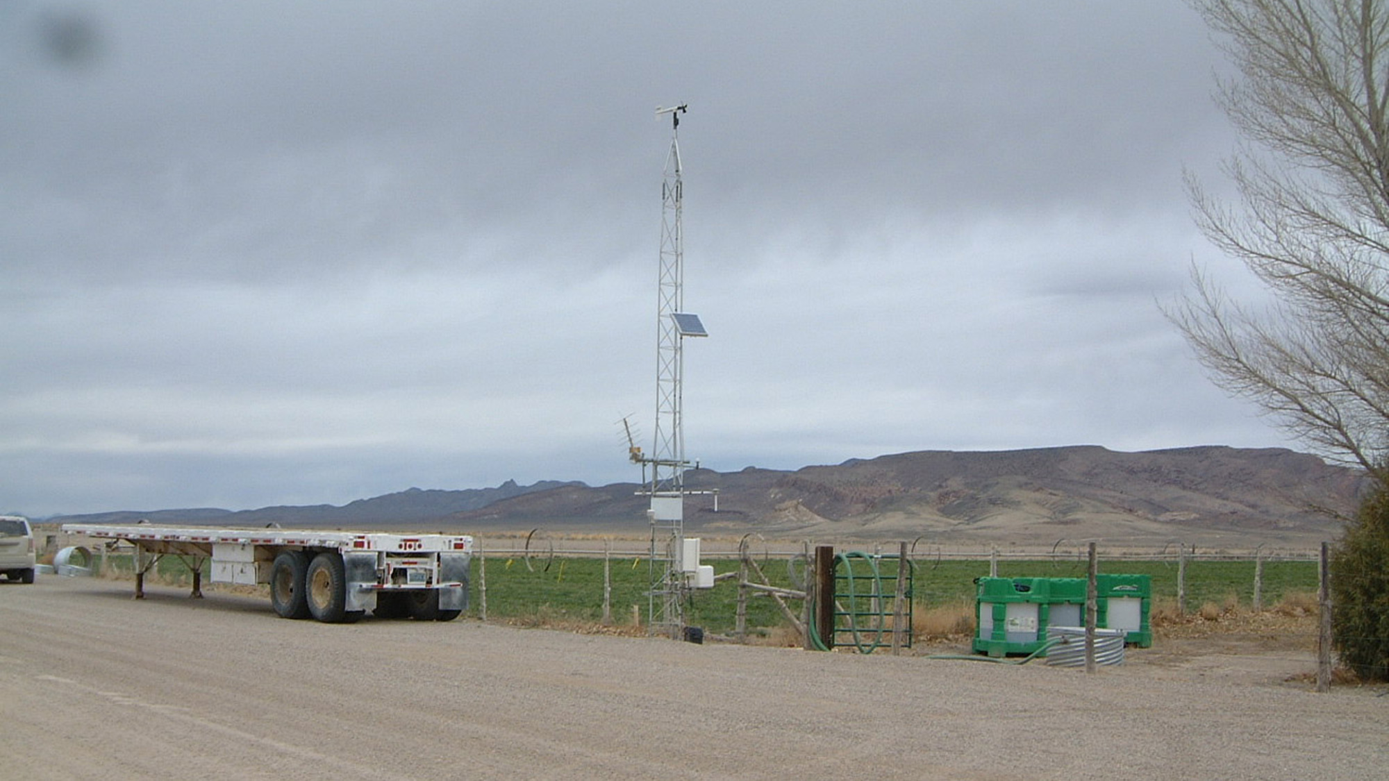 A weather monitoring station with sensors and a solar panel is installed near a road and a flatbed trailer, with mountains and fields in the background.