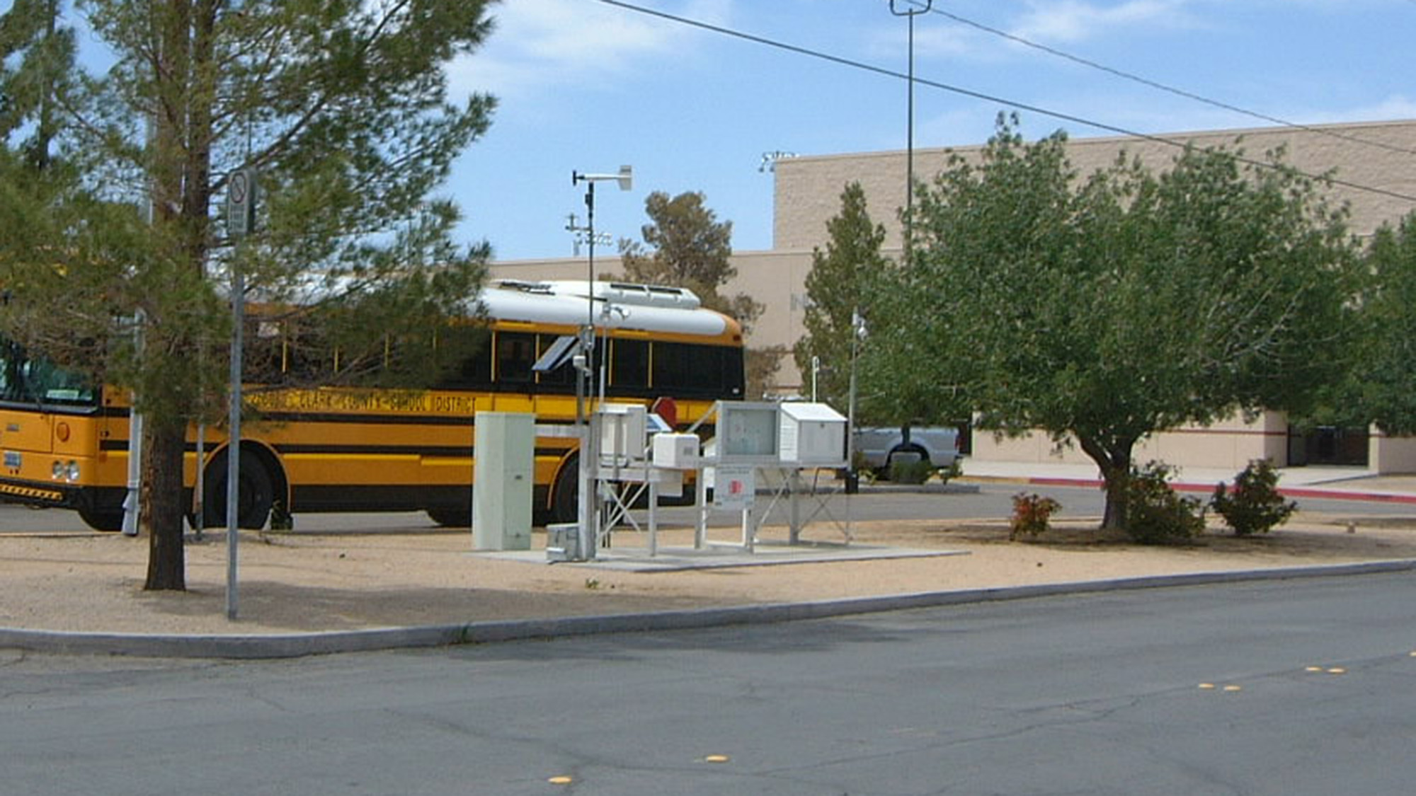 A yellow school bus is parked near a weather monitoring station, trees, and a beige building on a partly cloudy day.