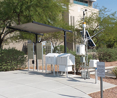 Las_Vegas_Nevada_Air_Monitoring_Thumbnail Outdoor weather monitoring station with multiple white instrument shelters, solar panels, sensors, informational signs, and nearby vegetation on a paved surface next to a building.
