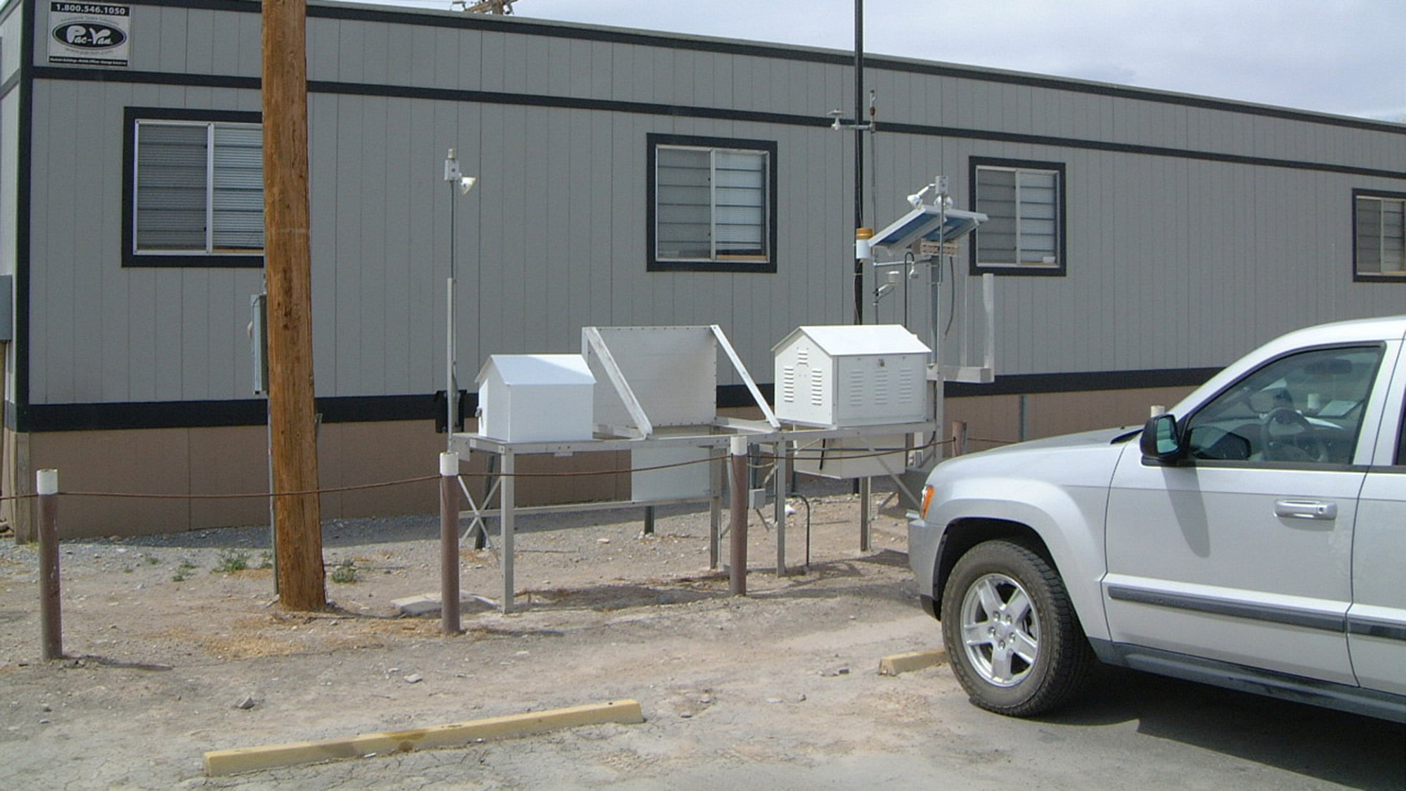 A white vehicle is parked beside a set of weather monitoring instruments installed on metal stands outside a grey portable building.