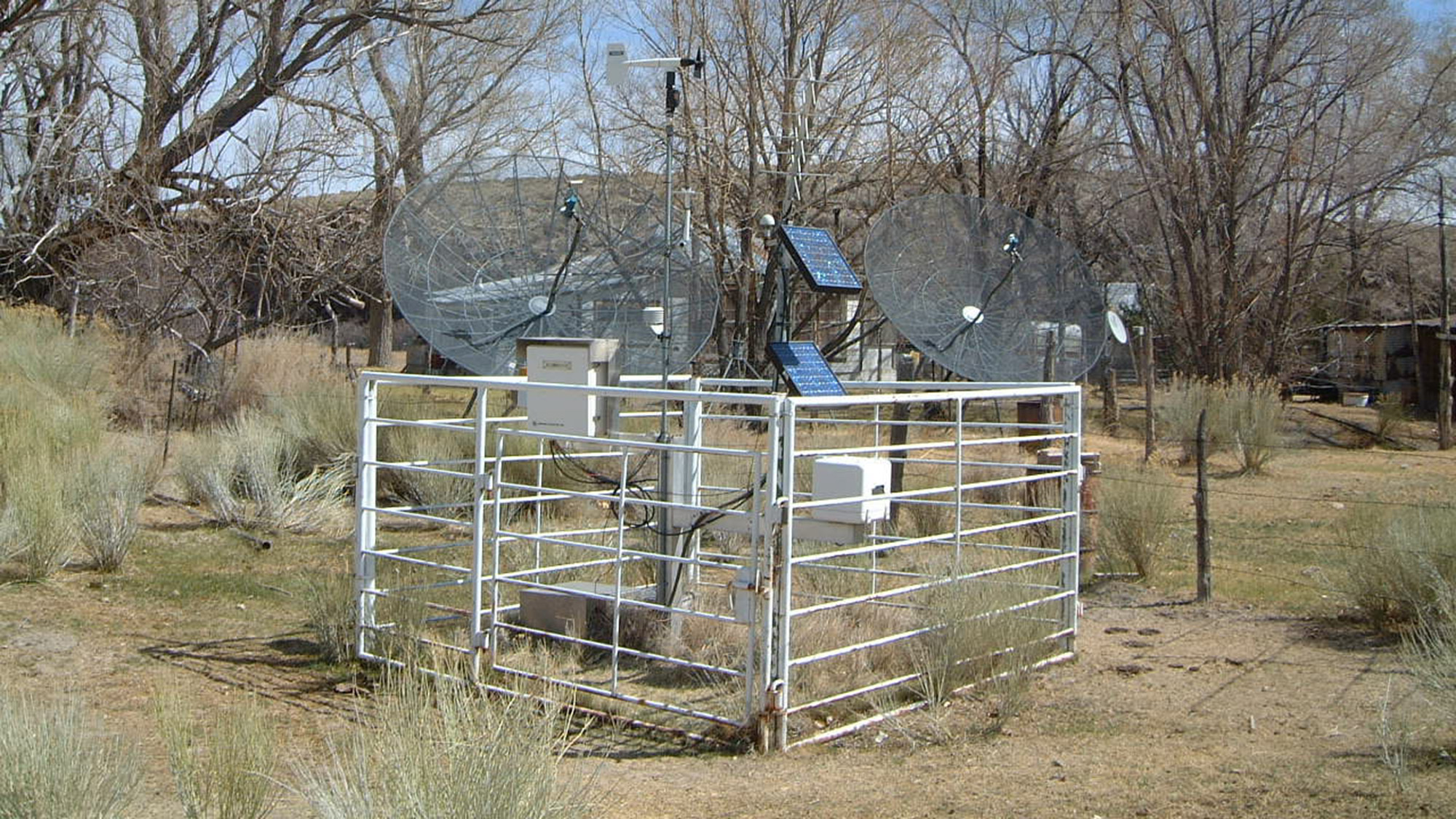 A fenced enclosure containing electronic equipment, solar panels, and two large satellite dishes set in a dry, grassy area with leafless trees in the background.