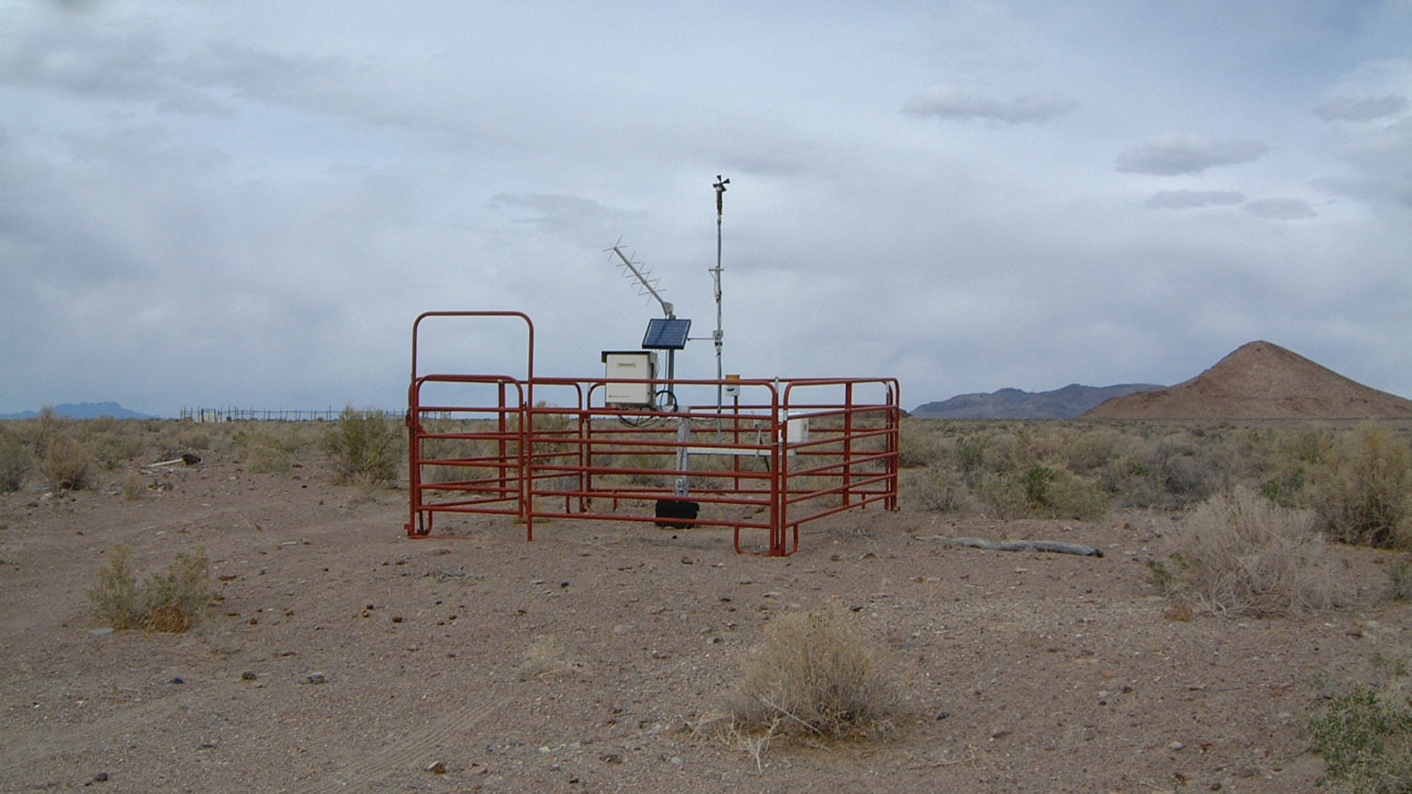 A weather monitoring station with solar panels and instruments is enclosed by a metal fence in a dry, desert landscape with distant hills.